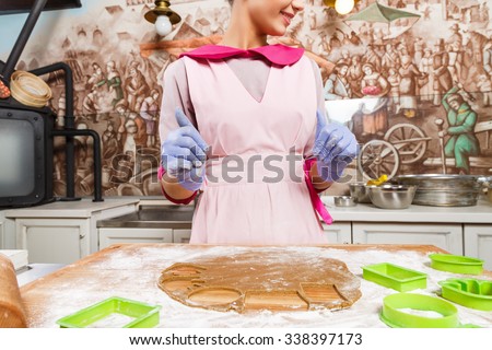 Young confectioner, wearing in pink dress, apron and violet gloves, cutting cookies from brown dough with green molds, on wooden board with rolling pin, in the kitchen, waist up