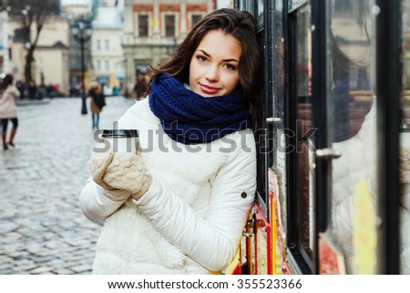Pretty young woman, with curly long hair, wearing in white coat, blue scarf and gloves, posing with cup of coffee near the tram, on the street of old European city, waist up