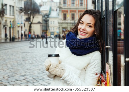 Smiling girl, with curly long hair, wearing in white coat, blue scarf and gloves, holding cup of coffee near the tram, on the street of old European city, waist up
