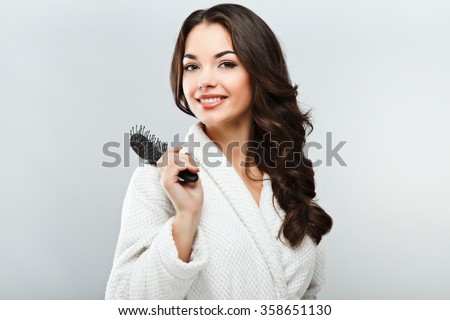Cute girl with dark hair, big eyes and dark eyebrows wearing white bath robe and holding hair brush, looking at camera and smiling, a model with light nude make-up, gray background, beauty photo