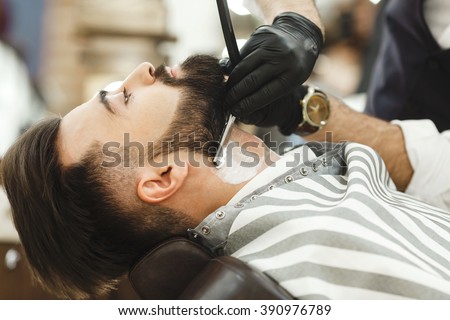Man\'s hands wearing black gloves and watch making a beard form with razor for man with dark hair and beard at barber shop, portrait, close up.