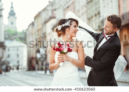 Wedding photo shooting. Bride and bridegroom walking in the city, looking at each other, holding bouquet and smiling. Groom holding veil of bride. Outdoor, cobbled street