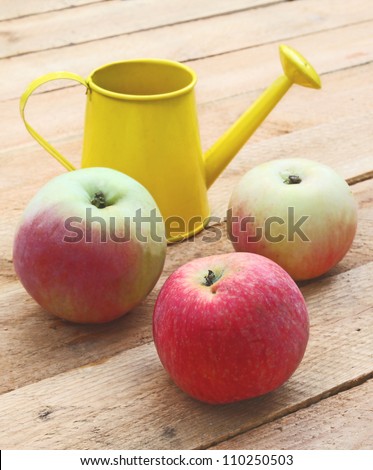 Three fruit of apple-tree and yellow watering-can on a wooden surface