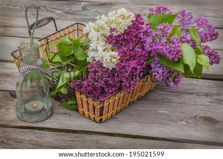 Basket with a bouquet of lilacs and light with a burning candle in the evening on a wooden table