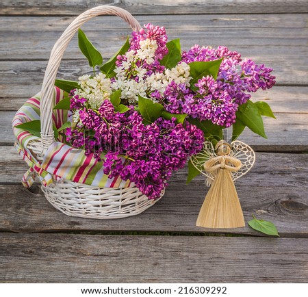 White basket with a branch of lilac and angel made of straw on a wooden table on a sunny spring day