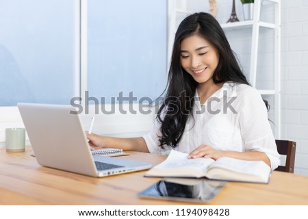 Education study abroad,Asian student girl sitting at table looking book while do homework with laptop making video call abroad using internet friend connection, happy mood smiling broadly in library
