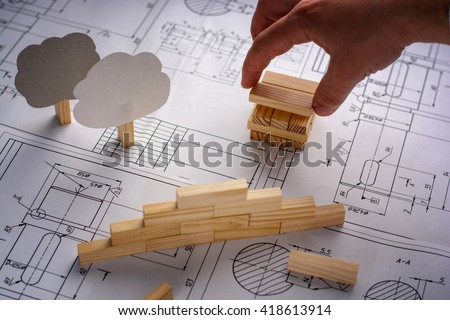 Man architect draws a plan, graph, design, geometric shapes by pencil on large sheet of paper at office desk and builds model house from wooden blocks (bars). Soft focus