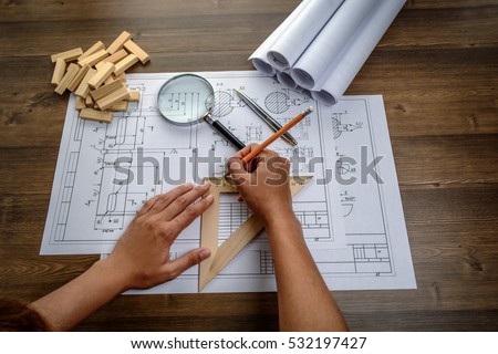 Girl architect draws a plan, graph, design, geometric shapes by pencil on large sheet of paper at office desk and builds model house from wooden blocks (bars)