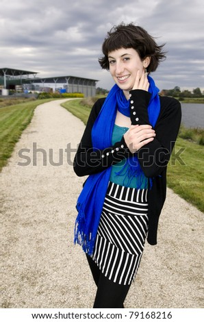 Attractive young woman with windblown hair wearing striped skirt and blue scarf in dynamic pose smiling at camera.