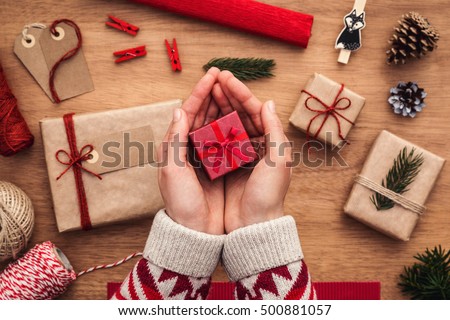 Woman holding small, red, christmas gift box over wooden desk with presents and decorations.