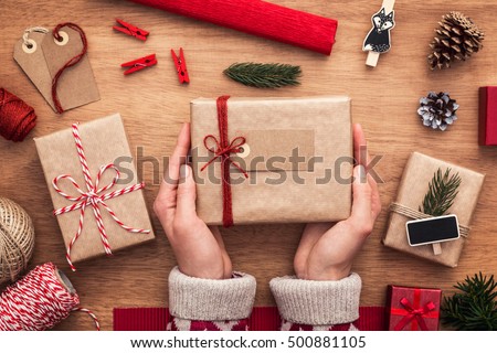 Christmas gifts wrapping ideas. Packing perfect presents. Close up on female hands. Wooden desk with decorative stuff: papers, ribbon, string, cones, labels and other things.