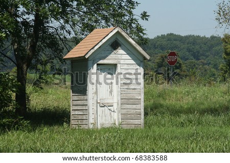 Old outhouse under the tree with a stop sign in the background seemingly reminding passers by that now might be a good time for a \