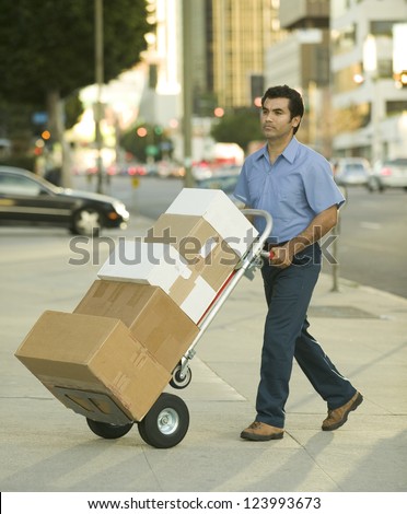 Hispanic delivery man walking on city sidewalk with hand truck full of boxes to deliver