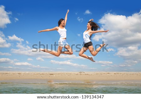 Jump up of two joyful young women on sea beach against blue sky