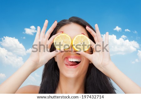 Portrait of joyful young woman holding lemon slices in front of eyes against blue sky