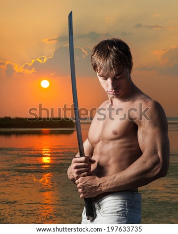 Muscular young man posing with Japanese sword on sunset background