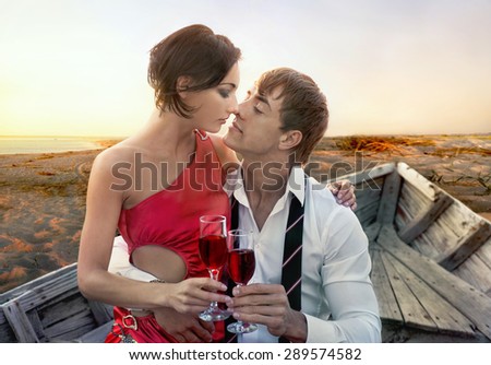 Portrait of romantic couple in love with wineglasses of red wine in old boat on sunset