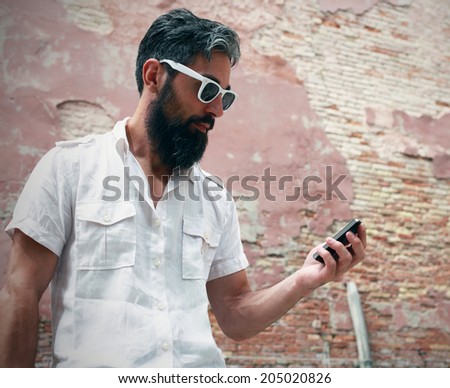 Profile of a hipster wearing white glasses with his smart phone, old wall on the background
