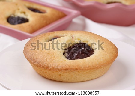 Small plum cake on a white cake stand. Plum cakes in pink pottery cake tins out of focus in the background.