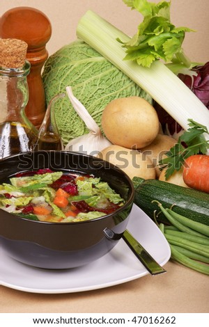 Vegetable soup (minestrone) in a black bowl and the organic seasonal vegetables used to prepare it. Studio shot over a light brown background.