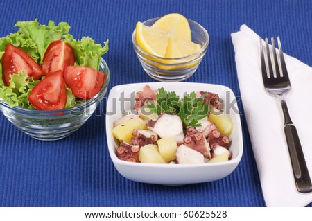 Laid table: boiled octopus with potatoes in a small white bowl, sliced lemon and mixed salad in two glass bowls. Studio shot