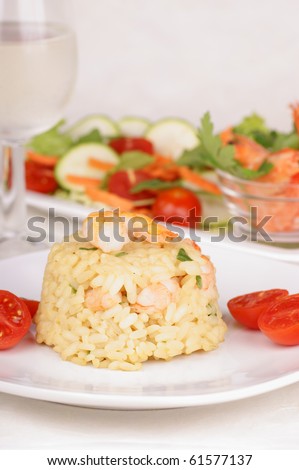Risotto with shrimps served on a white plate and decorated with cherry tomatoes. Shrimps, mixed salad and a glass of white wine in the background. Selective focus, extremely shallow DOF.