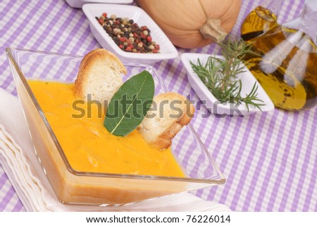 Cream of squash soup in a squared glass plate with toasted bread and a bay leaf. Pumpkin, whole peppercorns, rosemary and  a cruet out of focus in the background.