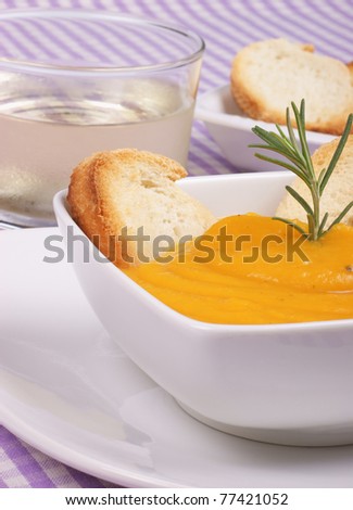 Cream of squash soup in a white plate with toasted bread and rosemary. A glass of wine and toasted bread out of focus in the background. Selective focus, shallow DOF.
