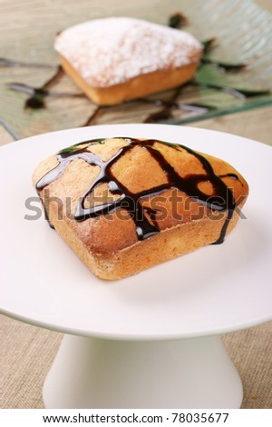 A small heart-shaped cake with chocolate topping served on a cake stand. A small cake with icing sugar served on a glass plate out of focus in the background