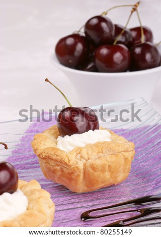 Small cherry tarts with whipped cream served on a glass plate. Cherries out of focus in the background. Selective focus, shallow DOF.