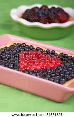 Tart with redcurrants and blueberries in a pink pottery cake tin. A tart with blueberries and raspberries out of focus in the background. Selective focus, shallow DOF.