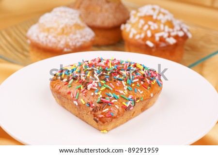 Small heart-shaped cake decorated with coloured sugar grains served on a white cake stand. Assorted muffins on a glass plate are out of focus in the background. Selective focus, shallow DOF