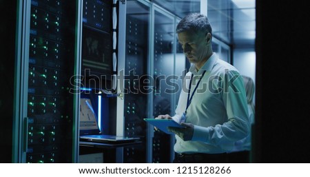 Medium shot of technician working on a tablet in a data center full of rack servers running diagnostics and maintenance on the system