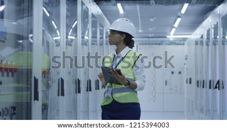 Medium shot of a female electrical worker reading her tablet and inspecting equipment in the control room of an electrical station