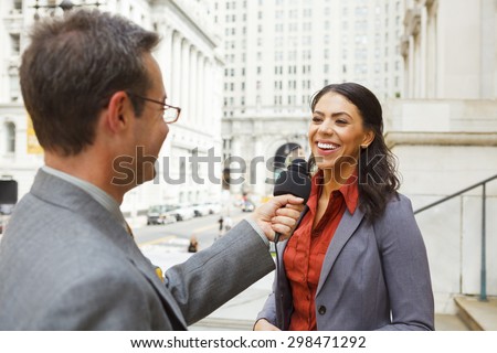A man with a microphone interviewing a smiling professionally dressed woman on the steps of a building in the city.