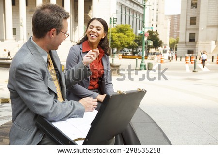 Professionally dressed man and woman laughing and conversing outdoors in a city environment.