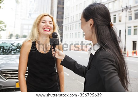A reporter with a microphone interviews a laughing woman. Focus is on blond woman's face.