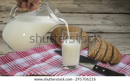 hand pouring milk from a jug into a glass on a wooden background grain bread on a napkin and knife
