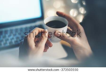 In female hands holding a cup of coffee or tea in front of a blue clean computer monitor on the background bokeh and light illuminations closeup