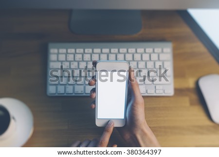 Closeup of a man pointing finger to smartphone with a blank screen monitor in the hands, working behind a computer keyboard and blue monitor on wooden table in the office horizontal