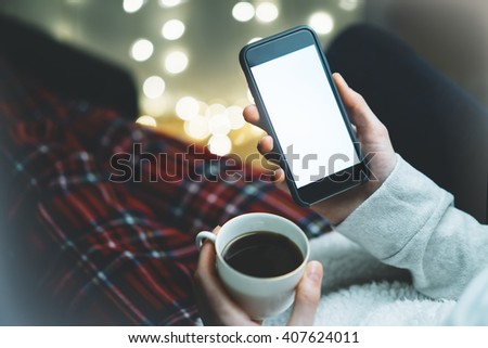 Girl holding smartphone with a empty blank screen monitor and a cup of coffee or tea on the background in a home atmosphere, hipster using in hands a smart phone with space for information, blur