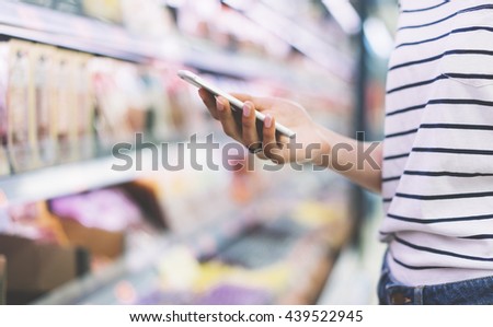 Hipster at grocery using smartphone. Young woman shopping healthy food in supermarket blur background. Close up view girl buy products using smartphone in store. Person comparing the price of produce