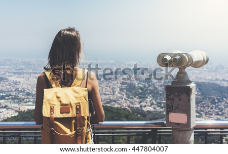 Hipster young girl with bright backpack looking on observation deck, tourist traveler on background panoramic view of the city, coin operated binoculars. Mock up for text message. Barcelona Tibidabo