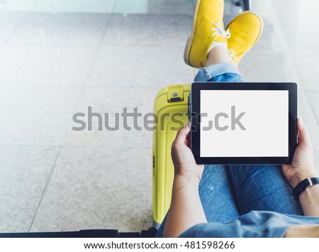 Young hipster girl at airport in yellow boot on suitcase waiting air flight, female hands holding computer in terminal departure lounge gate, traveler trip concept, mock up of blank screen tablet