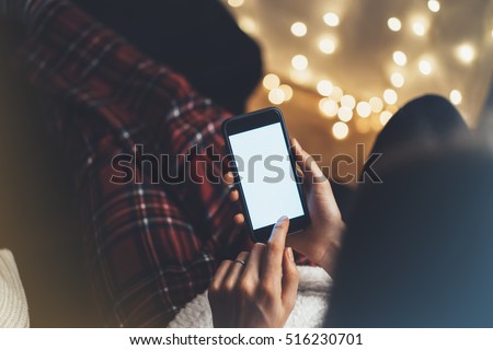 Hipster girl using phone in a home atmosphere, person holding smartphone on background glow bokeh Christmas illumination, female hands texting on relax glitter xmas decoration, mockup templates, blur