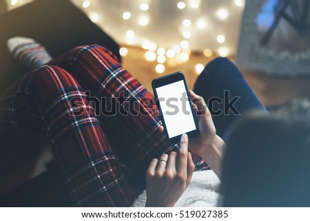 Hipster girl using mobile phone in a home atmosphere, person holding smartphone on background glow bokeh Christmas illumination,  hands texting on relax glitter xmas decoration, mockup templates, blur