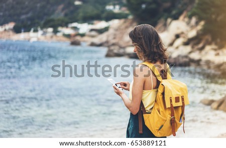 Hipster girl with backpack hold on smart phone gadget in sand coastline, mock up. Traveler using in female hand mobile on background beach seascape. Tourist look on blue sun ocean, summer lifestyle