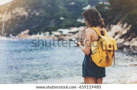 Hipster girl with backpack hold on smart phone gadget in sand coastline. Traveler using in female hand mobile on background beach seascape horizon. Tourist look on blue sun ocean, summer lifestyle