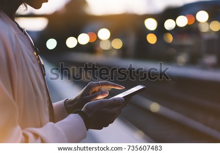 Woman waiting on station platform on background light train using smart phone in night. Tourist texting message and plan route of stop railway, railroad transport, booked. Enjoying travel concept
