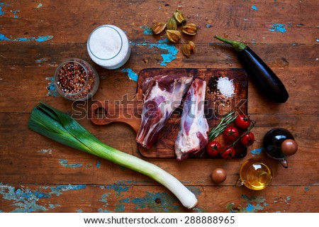 on the cutting board is fresh mutton shank is close eggplant fresh and tomatoes,bottle with oil and soy sauce are next to a cutting board, Cook rubbed in lamb ?oarse sea salt and pepper